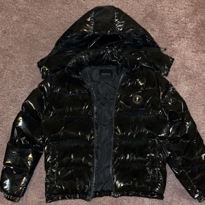Svart shiny pufferjacka från Trapstar - Säljer en äkta svart, glansig pufferjacka från Trapstar i storlek M. Ända negativa är att dragkedjan till luvan är trasig,  innerfickan är också trasig Kvitto finns!