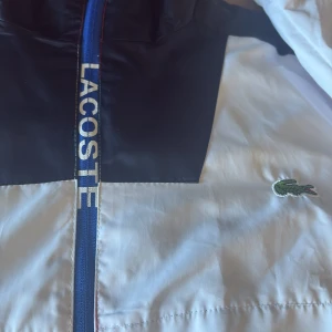 Färgstark vindjacka från Lacoste - Snygg vindjacka från Lacoste med cool färgblock-design i vitt, marinblått och rött. Jackan har huva, dragkedja med Lacoste-tryck och klassisk krokodillogga på bröstet. Stora Lacoste-bokstäver på ryggen. Perfekt för dig som gillar sportig och trendig stil.m