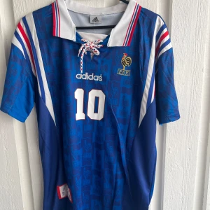 Frankrike Zidane 10 Adidas fotbollströja - Säljer en klassisk blå fransk landslagströja från Adidas med nummer 10 och Zidane på ryggen. Tröjan har vita och röda detaljer, snörning vid kragen och broderad FFF-logga. Kortärmad med mönster av tuppar och FFF-emblem i tyget. Perfekt för dig som älskar fotbollshistoria.