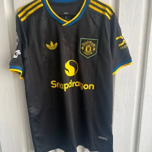 Manchester United svart matchtröja Mainoo - Snygg svart Manchester United fotbollströja från Adidas med gula och blå detaljer, korta ärmar och tryck 'Mainoo 37' på ryggen. Tröjan har klubbmärke, sponsorlogga och Champions League-märke på ärmen. Tillverkad i lätt, ventilerande material.