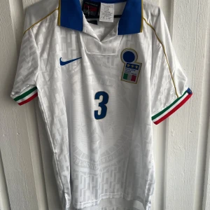 Italien Maldini #3 Nike fotbollströja - Snygg vit Italien fotbollströja från Nike med blå krage, guldiga detaljer och nummer 3 på bröstet och ryggen. MALDINI står tryckt på ryggen. Ärmarna har italienska flaggans färger och ett subtilt geometriskt mönster över hela tröjan. Perfekt för dig som älskar klassisk fotbollsstil.