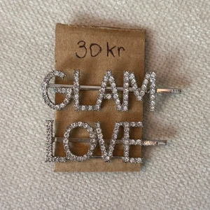 Glittriga hårspännen med text - Två hårspännen i silverfärg med gnistrande stenar som bildar orden GLAM och LOVE. Perfekta för att ge din frisyr extra bling och attityd. Stora bokstäver och riktigt iögonfallande design.