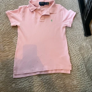Rosa Polo Ralph lauren piké - Snygg rosa t-shirt från Polo Ralph Lauren i slim fit med klassisk krage och korta ärmar. T-shirten har en liten grön logga broderad på bröstet och är gjord i mjuk bomull. Perfekt för dig som gillar stilrena och ikoniska plagg.Med lite svarta färgprickar som man kan säkert få bort i en tvätt