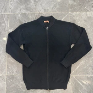 Svart stickad kofta med dragkedja - Svart stickad kofta från Raggen med hel dragkedja framtill och ribbade muddar vid ärmslut och nederkant. Klassisk och enkel design som passar till många olika stilar. Perfekt för kyliga dagar och enkel att matcha med jeans eller chinos.