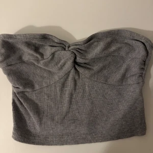 Grå ribbad tubtopp - Säljer en grå bandeau tubtopp med snygg twistad detalj framtill. Toppen är ribbad och har en mjuk, stretchig känsla som sitter skönt mot kroppen. Perfekt för dig som gillar minimalistisk stil och vill ha en enkel men trendig topp till din garderob.