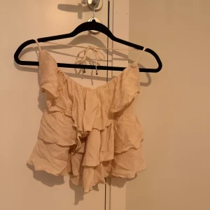 Beige volang topp med knytband - Supersöt beige halterneck topp med volanger och tunna axelband som knyts i nacken. Toppen har en luftig och flowy passform, perfekt för varma dagar. Materialet är lätt och känns skönt mot huden. Aldrig använd!