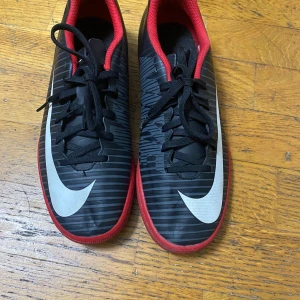 Nike MercurialX svarta fotbollsskor - Nike MercurialX fotbollsskor i svart med röda detaljer och innersula. Skorna har ett randigt mönster, stor vit swoosh på sidan och snörning framtill. Platt röd sula och låg profil gör dem perfekta för inomhusspel. Snygg design som sticker ut på planen.