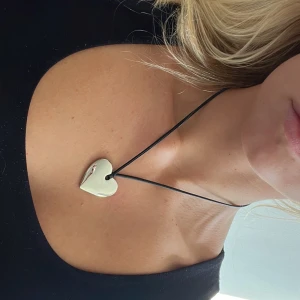 Halsband med silverfärgat hjärta - Snyggt halsband med ett blankt, silverfärgat hjärta som hänge. Hängs på en svart, tunn rem med metallspänne. Perfekt för dig som gillar stilrena och romantiska detaljer. Hjärtat har en mjuk, rundad form och ger en cool vibe till din outfit.