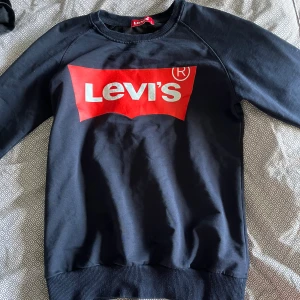 Svart Levi's sweatshirt med röd logga - Snygg svart sweatshirt från Levi's i storlek L med den klassiska röda loggan tryckt på bröstet. Tröjan har rund hals, långa ärmar och är gjord i mjuk bomull. Tröjan är i L men jag som är S tycker att den sitter bra!