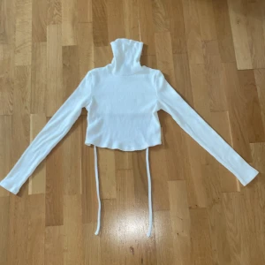 Vit ribbad croppad polotröja - Säljer en vit croppad polotröja med ribbad struktur och långa ärmar. Tröjan har hög polokrage och snörning i midjan som ger en snygg detalj. Perfekt att styla med jeans eller kjol för en trendig look.