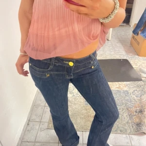 Mörkblå bootcut jeans med gula detaljer - Snygga mörkblå jeans med bootcut passform och låg midja. Jeansen har coola gula knappar och nitar, samt kontrastsömmar som ger en unik look. Perfekta för dig som gillar en klassisk men ändå trendig stil. Materialet är klassisk jeans, vilket gör dem både bekväma och slitstarka.