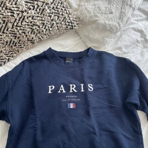 Mörkblå sweatshirt med Paris-tryck - Snygg mörkblå sweatshirt från Gina Tricot med stort vitt Paris-tryck och fransk flagga på bröstet. Tröjan har rund hals, långa ärmar och en relaxed passform. Perfekt för dig som gillar streetstyle och vill ha en chill vibe.