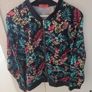 Blommig bomberjacka från Active Wear Happy Holly. storlek 44/46 XL - Färgglad bomberjacka med blommigt mönster i turkos, rosa, gul och röd på svart botten. Jackan har dragkedja framtill och ribbade muddar vid ärmslut och nederkant. Storlek 44/46 XL.Från rök och djurfritt hem.