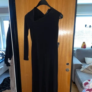Svart heldress från Nelly - Snygg svart heldress från Nelly med asymmetrisk design och oneshoulder. Ena sidan har lång ärm medan den andra är ärmlös, vilket ger en cool och modern look. Klänningen är figurnära upptill och har ett mjukt, stretchigt material som sitter bekvämt.