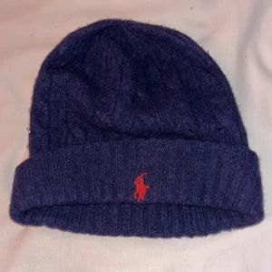 Mörkblå ullmössa från Polo Ralph Lauren - Snygg mörkblå mössa från Polo Ralph Lauren i mjuk ull. Klassisk ribbad kant och ikonisk röd broderad logga framtill. Perfekt för kalla dagar och ger en clean look med lite lyxig känsla. Passar dig som gillar stilrena accessoarer.