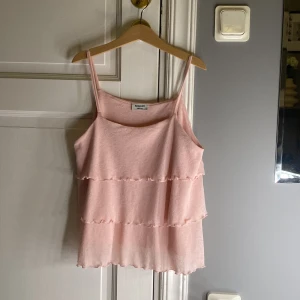 Rosa volang-linnetopp från Kappahl - Supersöt ljusrosa linnetopp med dubbla volanger och tunna axelband från Kappahl. Toppen har en flowy passform och är tillverkad i mjuk bomull, perfekt för varma dagar. Volangdetaljerna ger en extra girly vibe och toppen är ärmlös för en chill look.