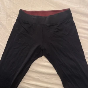 Svarta leggings från Newbody - Svarta leggings från Newbody med bred, mörkröd linning som ger extra komfort. Snyggt diskret logotyptryck längs benet. Stretchigt material som sitter tight och följer kroppen, perfekt för träning eller chill. Passar dig som gillar enkel och stilren look.