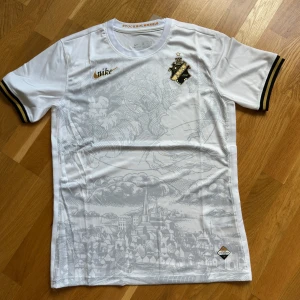 Vit AIK fotbollströja från Nike - AIK fotbollströja i vitt med grått stadsmotiv på framsidan, guldiga detaljer och klubbmärke på bröstet. Svarta och gula ränder på ärmsluten ger extra stil. Tillverkad i lätt och ventilerande material, perfekt för match eller träning.