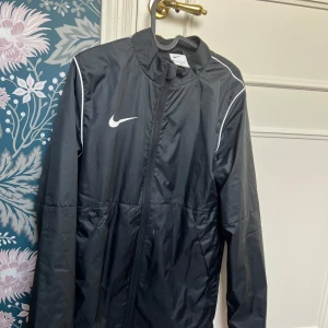 Svart Nike vindjacka med dragkedja - Svart vindjacka från Nike med vit logga på bröstet och vita detaljer längs axlarna. Jackan har hel dragkedja framtill, ståkrage och långärmad design. Tillverkad i lätt polyester som passar perfekt för aktiva dagar och sport.