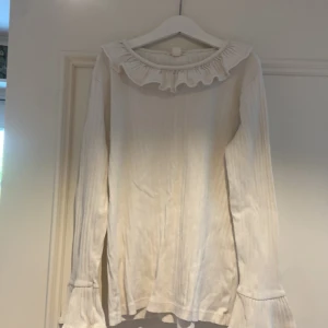 Vit ribbad blus med volang från Crewcuts - Supersöt vit ribbad blus från Crewcuts med volang runt halsen och vid ärmsluten. Långärmad och i mjukt material som känns skönt mot huden. Perfekt för dig som gillar en clean och feminin stil med lite extra detaljer.