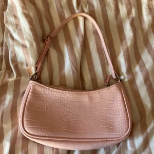 Ljusrosa axelväska från Monki - Säljer en ljusrosa axelväska från Monki med croco-mönstrad yta i syntetmaterial. Väskan har en justerbar axelrem och dragkedja upptill. Perfekt storlek för det viktigaste och har en snygg, modern form som ger en cool vibe.