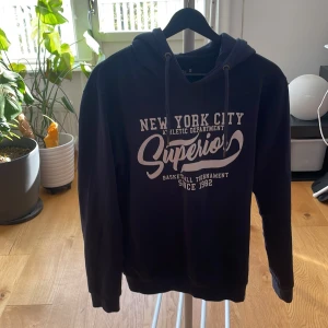 Mörkblå hoodie från Indicode - Snygg mörkblå hoodie från Indicode med vit tryckt text 'New York City Superior' på bröstet. Tröjan har huva med snörning och långa armar.
