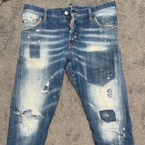 Dsquared2 blå jeans med slitningar - Säljer ett par blå Dsquared2 jeans med coola slitningar, patchade hål och färgstänk i vitt och grönt. Modellen har normal passform, klassisk femficksdesign och tydliga blekta partier. Jeansmaterialet är bomull och de har en snygg streetstil.