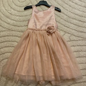 Champagnefärgad glittrig klänning H&M - Superfin champagnefärgad klänning från H&M med glittrigt liv och voluminös kjol i tyll. Klänningen har ärmlös design, rund hals och en dekorativ blomma i midjan. Baksidan har knappar för enkel påklädning. Perfekt för speciella tillfällen!