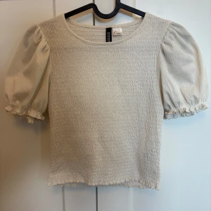 Krämvit blus med puffärm från Divided - Supersöt blus i krämvit färg från Divided, med puffiga korta ärmar och smockad struktur över hela kroppen. Rund halsringning och små volangdetaljer vid ärmslut och nederkant.  