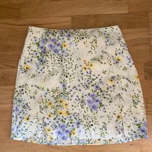 Blommig kortkjol från Pimkie - Superfin kortkjol från Pimkie med blommigt mönster i lila, gul och grönt på vit botten. Kjolen har en rak passform och dragkedja bak. Perfekt för dig som vill ha en söt och somrig vibe i garderoben!