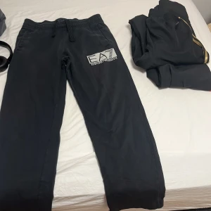 Svarta mjukisbyxor EA7 Emporio Armani - Svarta mjukisbyxor från EA7 Emporio Armani med stort logotryck på vänster ben. Byxorna har elastisk midja med snörning och sidofickor. Perfekta för chill eller träning, riktigt snygga och bekväma med loose passform.