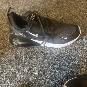 Nike Air Max 270 svarta sneakers - Nike Air Max 270 sneakers i svart med vita detaljer och synlig Air-enhet i hälen. Snygg mesh-överdel med ventilerande struktur, klassisk snörning och rund tå. Sulan är vit med svart och ger en sportig vibe. Perfekt för dig som gillar streetstyle och komfort.