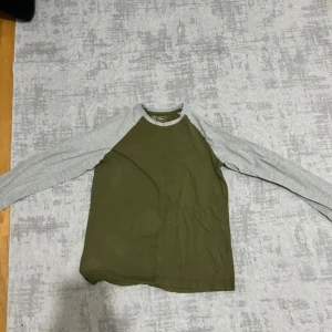 Grön långärmad tröja från Friboo - Säljer en stilren långärmad tröja från Friboo i storlek 158/164. Tröjan har olivgrön kropp och ljusgrå ärmar med rund halsringning. Perfekt för dig som gillar enkel och sportig stil. Materialet är mjuk bomull som känns skönt mot huden.