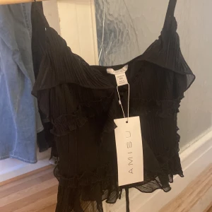 Svart volangig linnetopp från Amisu - Supersnygg svart linnetopp från Amisu med tunna axelband och volangdetaljer. Toppen är plisserad och har en lätt transparent känsla som ger extra edge. Perfekt att styla med jeans eller kjol för en cool look. det är helt ny med prislappen kvar