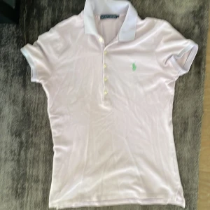 Lila pikétopp från Ralph Lauren - Snygg lila pikétopp från Ralph Lauren med klassisk krage och korta ärmar. Toppens framsida har fyra vita knappar och den ikoniska gröna polospelaren broderad på bröstet. Perfekt för en chill och sportig look.