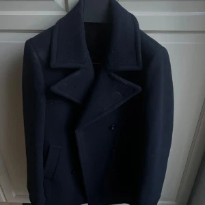 Mörkblå skepparkavaj från Suitsupply - Snygg mörkblå skepparkavaj från Suitsupply i ren ull. Jackan har breda slag, dubbelknäppning och två framfickor. Baksidan har ett dekorativt band med knappar. Perfekt för dig som vill ha en klassisk och stilren look. Nypris 4500