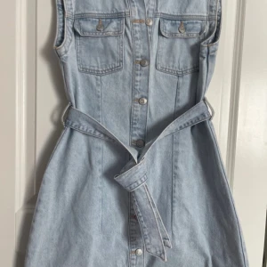 Ärmlös jeansklänning från Zara - Snygg ljusblå jeansklänning från Zara med krage, knappar hela vägen framtill och två bröstfickor med lock. Klänningen är ärmlös och har ett matchande bälte i midjan för extra stilpoäng. Perfekt för dig som gillar en cool och avslappnad look. Använd 1 gång 