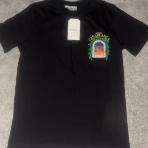 Svart Casablanca t-shirt med tryck - Svart t-shirt från Casablanca med färgstarkt grafiskt tryck fram och bak, där det står 'Avenida Casablanca' och motiv av en byggnad i en fönsterliknande ram med gröna blad. Klassisk rund hals och korta ärmar. Skön bomullskvalitet och normal passform.