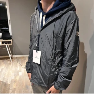 Grå moncler Windbreaker  - Snygg grå vindjacka från Moncler med huva och coola snörningsdetaljer. Jackan har dragkedja framtill och Moncler-logga på ärmen. På huvan finns en röd, vit och marinblå rand som ger extra stilpoäng. Perfekt för dig som gillar streetwear och vill ha något lätt och funktionellt.