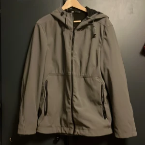 Grå vindjacka med huva från Zara - Grå vindjacka/regnjacka från Zara med snygg huva och svarta detaljer. Jackan har dragkedja framtill, två fickor med dragkedja och justerbar snörning i nederkant. Perfekt för blåsiga dagar och enkel att matcha med din stil.