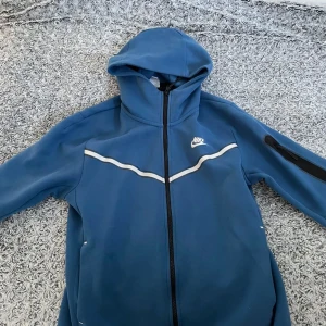 Blå Nike vindjacka med huva - Snygg blå vindjacka från Nike med huva och hel dragkedja framtill. Jackan har vita detaljer över bröstet och en praktisk ficka med dragkedja på ärmen. Perfekt för dig som gillar sportig stil och vill ha något bekvämt och funktionellt. Den är storlek M men passar S 
