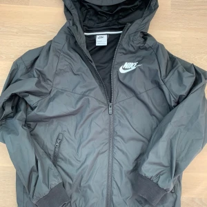 Svart vindjacka från Nike - Svart Nike vindjacka med huva och dragkedja framtill. Jackan har ribbade muddar, två sidofickor med dragkedja och ett Nike-logotryck på bröstet. Tillverkad i lätt polyester med meshfoder för extra komfort. Perfekt för dig som gillar sportig stil. Säljs för 350kr nypris 749kr, köparen står för frakten