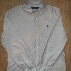 Grå skjorta från Ralph Lauren S - Snygg grå skjorta från Ralph Lauren i storlek S. Klassisk button-down krage och broderad marinblå logga på bröstet. Skjortan har långa ärmar och knappar framtill. Perfekt för dig som gillar stilrena och tidlösa plagg.