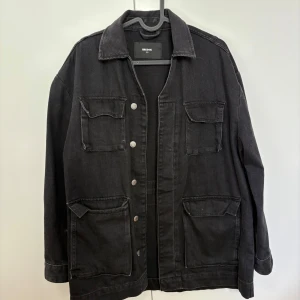 Svart jeansjacka från Bik Bok XS - Säljer en svart oversize jeansjacka från Bik Bok i storlek XS. Jackan har fyra stora fickor med lock framtill och silverfärgade knappar. Klassisk krage och rak passform, perfekt att slänga över en hoodie eller t-shirt. Materialet är robust denim och färgen är mörkt svart.