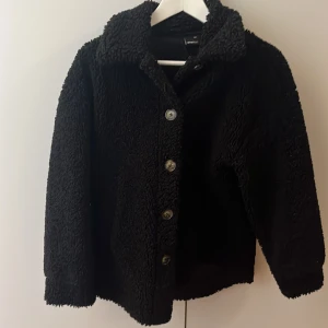 Svart teddyjacka från Gina Tricot - Mysig svart teddyjacka från Gina Tricot i storlek XXS men stor i modellen och upplevs mer som S/M Jackan har en fluffig struktur, klassisk krage och knäppning med fem knappar framtill. Perfekt för att hålla värmen och ge en cool vibe till din outfit.