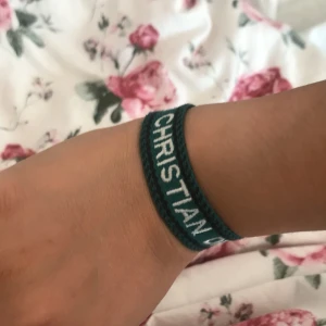 Grönt Christian Dior armband i tyg - Snyggt grönt armband från Christian Dior i vävt tyg med vit text. Armbandet har justerbar knytning med tofsar i ändarna och en cool, casual vibe. Perfekt accessoar för att lyfta din outfit med en touch av lyx och streetstyle.