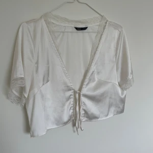 Vit satin crop top - Säljer en vit crop topp i satin från SHEIN. Toppen har korta ärmar med spetsdetaljer och knyt framtill. Djup v-ringning med spetskant ger en romantisk känsla. 
