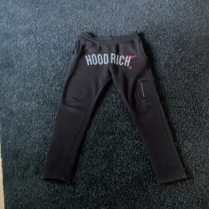 Svarta mjukisbyxor från Hoodrich - Svarta mjukisbyxor från Hoodrich med tryckt logga framtill i vitt och rött. Byxorna har en normal passform och resår i midjan. Perfekta för en avslappnad stil och har en ficka på sidan av benet.