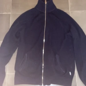 Mörkblå stickad jacka från Jack & Jones - Mörkblå stickad jacka från Jack & Jones med hel dragkedja framtill och hög krage. Jackan har två fickor och ribbade muddar vid ärmslut och nederkant. Perfekt för lager-på-lager under kyliga dagar.