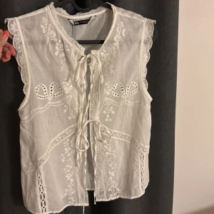 Vit broderad topp med knyt från Zara - Superfin vit topp från H&M med broderade detaljer och spets. Toppen har knyt framtill och vackra hålbroderier samt volangkant vid axlarna. Aldrig använd med prislappen kvar.